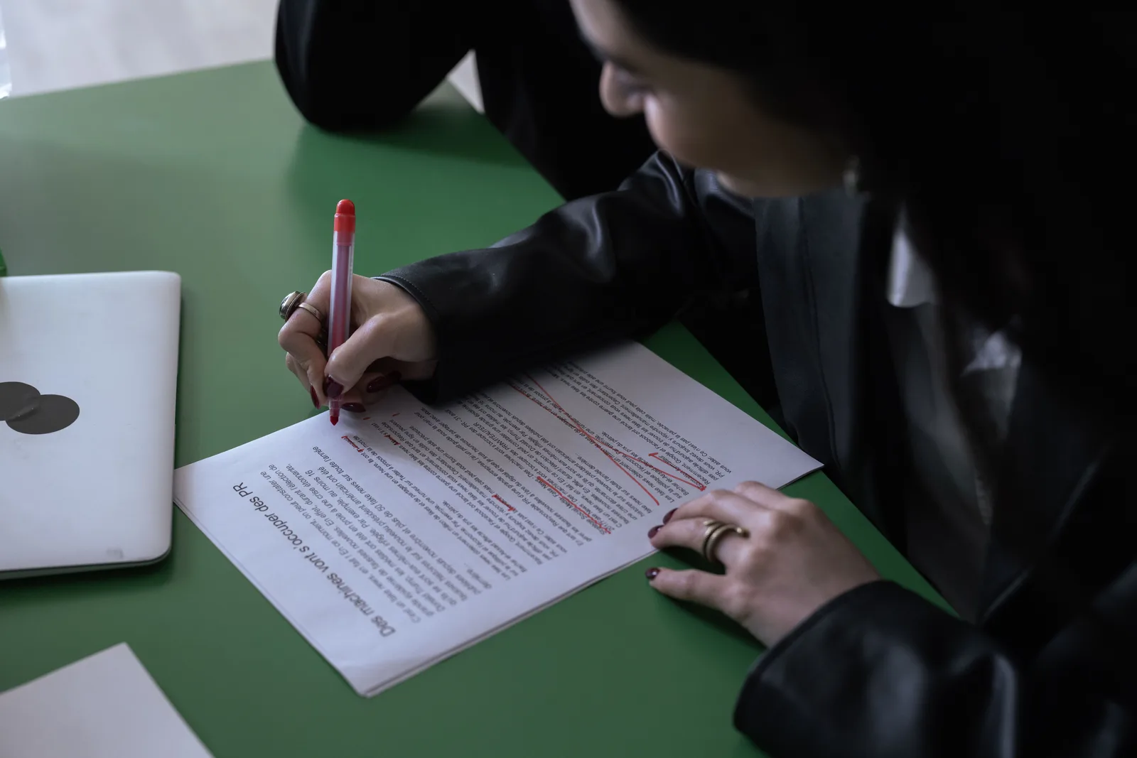 A woman in a suit uses a red pen to edit a printed document on a green table with a laptop. Legal marketing compliance examples with the reasoning laid bare