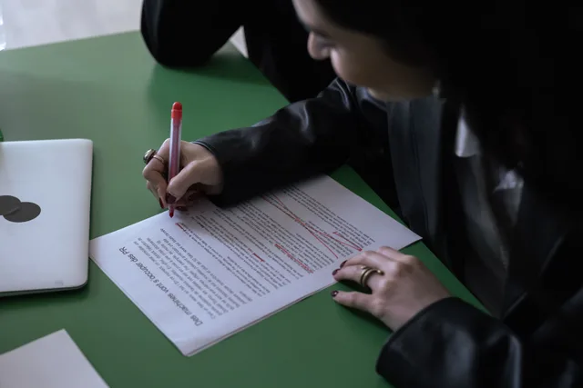A woman in a suit uses a red pen to edit a printed document on a green table with a laptop. Legal marketing compliance examples with the reasoning laid bare